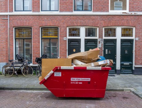 A large red skip bin positioned on the pavement in front of a brick residential building with three dark green front doors. The skip is filled with assorted household waste, including flattened cardboard boxes, a large rolled-up brown paper parcel, and various other debris, indicating a house clearance process typical of Bristol or surrounding areas. Bicycles are parked to the left of the skip, leaning against the building, with a metal railing nearby. The building features large, arched windows with decorative yellow detailing above each door, and the scene is set during daylight with overcast weather, contributing to a neutral, professional atmosphere appropriate for a rubbish removal service like House Clearance Brentwood, serving the Brentwood area and nearby postcode regions. The scene captures an organized, typical street scene where domestic waste collection or clearance activities are visibly underway, emphasizing the company's local expertise in household rubbish disposal particularly in the context of property cleanouts and waste management in the town.