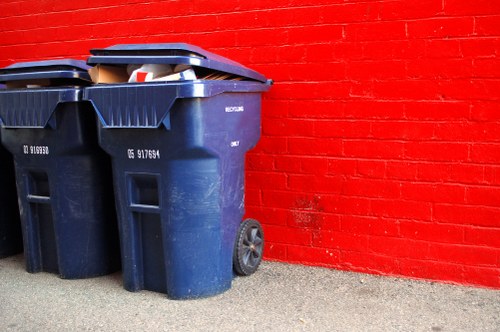 Two large dark blue plastic wheelie bins with black lids are positioned on a paved surface beside a bright red brick wall. One bin's lid is slightly open, revealing some visible rubbish inside, including cardboard and plastic packaging. The bins are situated close together, with one slightly in front of the other, and both have black wheels at their bases for mobility. The textured brick wall behind them features horizontal mortar lines, contrasting sharply with the smooth, worn concrete ground. The scene appears to be outdoors, possibly in a residential driveway or service area. This setting aligns with typical rubbish collection and waste disposal environments, frequently serviced by companies like House Clearance Brentwood, which specialise in rubbish removal services in the Brentwood area. The image captures the practical side of waste management, emphasizing the importance of proper bin storage and disposal in urban and suburban settings within the Brentwood postcode area.