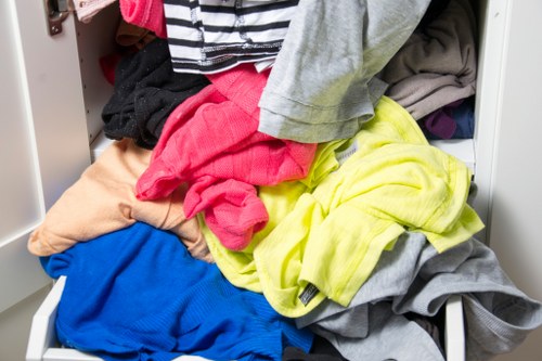 An open white metal wardrobe or storage cabinet with shelves containing a disorganized pile of mixed laundry and clothing items, including brightly colored t-shirts, trousers, and towels in shades of pink, yellow, blue, grey, and black. The garments appear crumpled and partially spilling out of the drawer, with some pieces draped over the edges. The environment suggests a domestic setting, likely a bedroom or laundry area, with visible metal shelf supports and a neutral background. This cluttered scene reflects typical rubbish or waste accumulation from household laundry, relevant to rubbish removal services provided by House Clearance Brentwood, in the context of clearing household waste or unwanted clothing items for disposal or recycling.