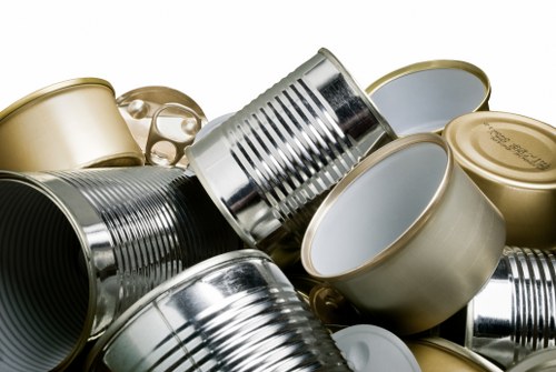 A close-up view of a collection of tin cans and metal food containers piled together, including silver cans with ribbed surfaces, and gold-toned, smooth-sided tins, some open and others with lids, set against a neutral background. The cans vary in size and shape, with some positioned horizontally and others tilted, displaying their open and closed tops, with visible ridges and reflective surfaces. The environment suggests an indoor setting, possibly part of a waste collection or recycling process, typical of rubbish removal services operating in areas like Brentwood. The image emphasizes the metallic textures and reflective qualities of the cans, reinforcing their material composition. This visual is relevant for services such as house clearance that involve clearing and disposing of household waste and recyclable materials as offered by House Clearance Brentwood, situated within the Brentwood area, which is associated with efficient rubbish removal and waste management. The lighting highlights the sheen and contours of the metal containers, contributing to a clear and detailed depiction suitable for accessibility and SEO relevance.